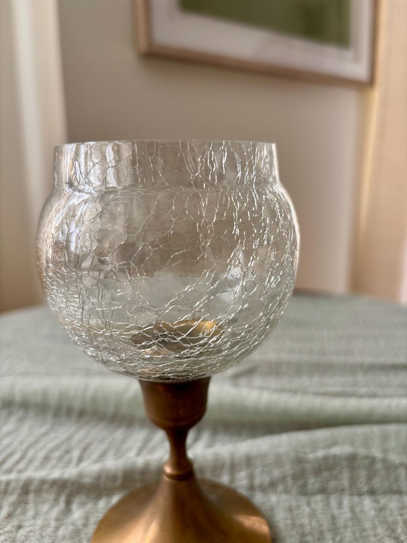 May include: A clear glass candle holder with a crackled texture, resting on a bronze-colored metal base. The candle holder has a rounded bowl shape and a slender stem, creating an elegant aesthetic.