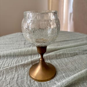 May include: A clear glass candle holder with a crackle finish bowl-shaped top. The holder is supported by a bronze-colored metal base and stem. The candle holder is designed to hold a small candle.