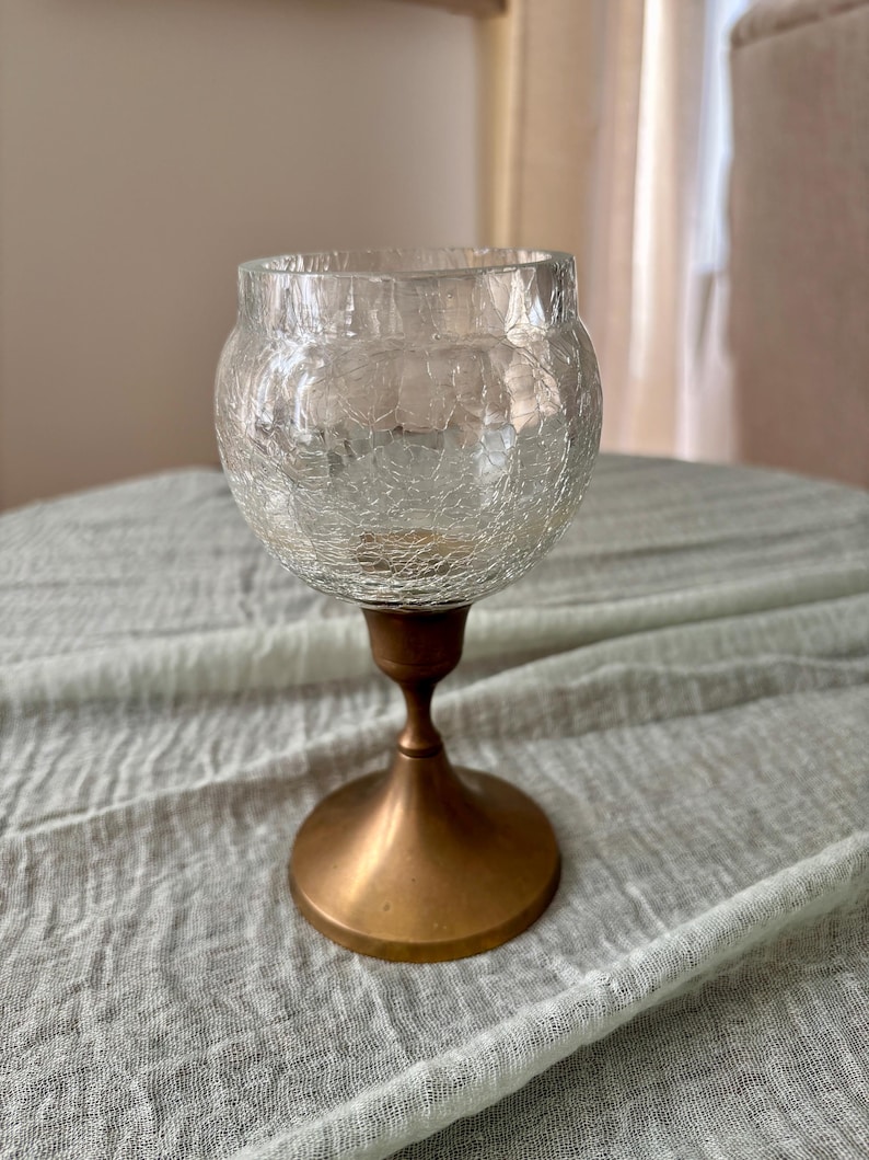 May include: A clear glass candle holder with a crackle finish bowl-shaped top. The holder is supported by a bronze-colored metal base and stem. The candle holder is designed to hold a small candle.