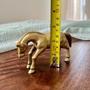 May include: A brass horse figurine, approximately 4 inches tall, stands on a wooden surface. The horse is in a trotting pose, with its head lowered. The figurine has a detailed mane and tail.