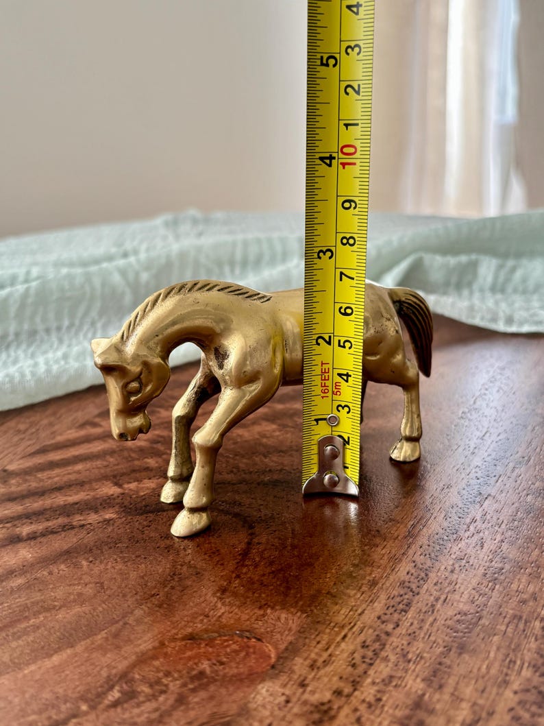 May include: A brass horse figurine, approximately 4 inches tall, stands on a wooden surface. The horse is in a trotting pose, with its head lowered. The figurine has a detailed mane and tail.