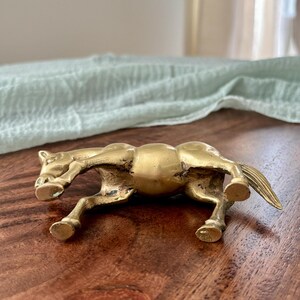 May include: A small, antique-style brass horse figurine in a running pose. The horse is detailed with visible muscles and a flowing mane and tail. The figurine is displayed on a dark wood surface.