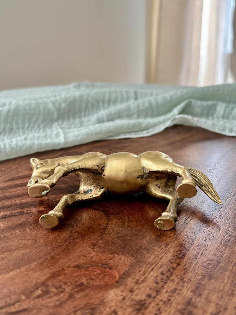 May include: A small, antique-style brass horse figurine in a running pose. The horse is detailed with visible muscles and a flowing mane and tail. The figurine is displayed on a dark wood surface.