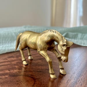 May include: A brass horse figurine in a trotting pose. The sculpture has a golden hue and is detailed with a mane, tail, and defined legs. The horse is positioned on a wooden surface, with a soft, light-colored background.