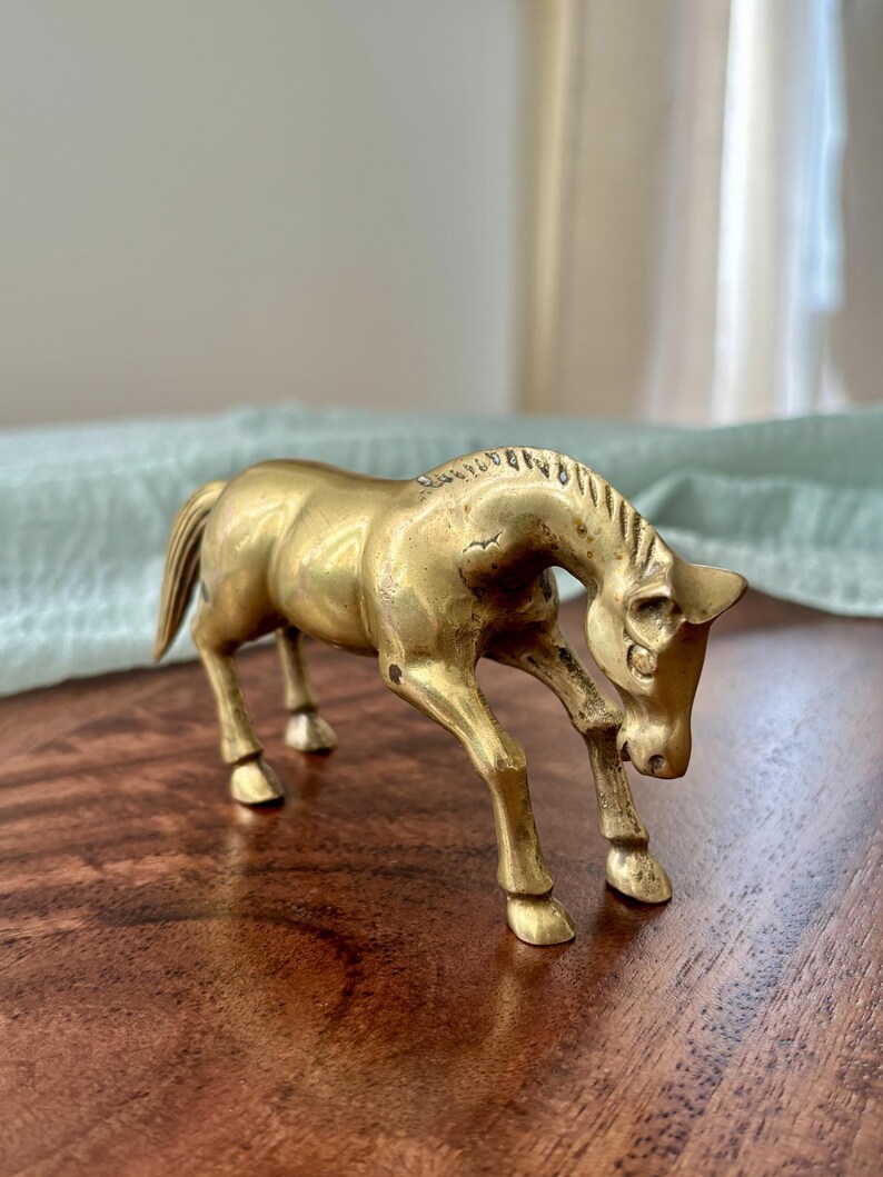 May include: A brass horse figurine in a trotting pose. The sculpture has a golden hue and is detailed with a mane, tail, and defined legs. The horse is positioned on a wooden surface, with a soft, light-colored background.