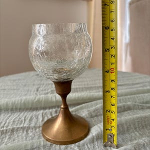 May include: A clear glass candle holder with a crackle finish, measuring approximately 6 inches tall. It features a bronze-colored metal base and stem. The candle holder is designed to hold a small candle and is suitable for home decor.