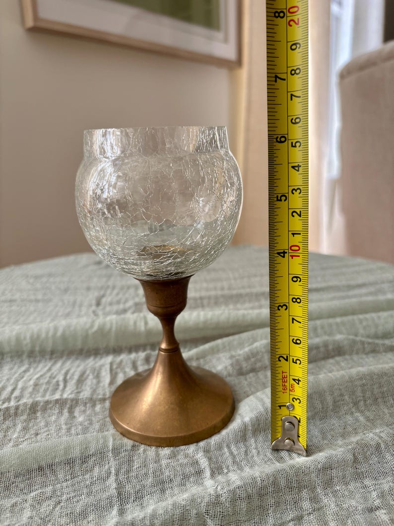 May include: A clear glass candle holder with a crackle finish, measuring approximately 6 inches tall. It features a bronze-colored metal base and stem. The candle holder is designed to hold a small candle and is suitable for home decor.