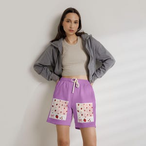 May include: A pair of lavender sweat shorts featuring two rectangular patches on the front. Each patch displays a white background with a pattern of red strawberries, pink bows, and light pink gingham stripes. The shorts have a white drawstring waist.