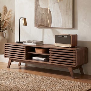 May include: A mid-century modern style wooden TV stand with slatted doors and angled legs. The stand has open shelving and a small speaker on top. A black and gold lamp and books are also on the stand. A beige rug is on the floor.