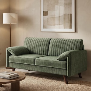 May include: A sage green velvet sofa with vertical channel tufting and two matching throw pillows. The sofa has dark wooden legs and is placed on a woven rug. A round wooden coffee table holds books and a vase with dried flowers.