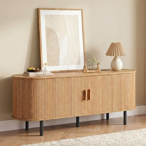 May include: A light wood-tone cabinet with a ribbed front and black legs. A framed abstract art print and a lamp with a pleated shade sit on top. The cabinet has two doors with vertical handles. A book with the text "CERENE UNION" is visible.