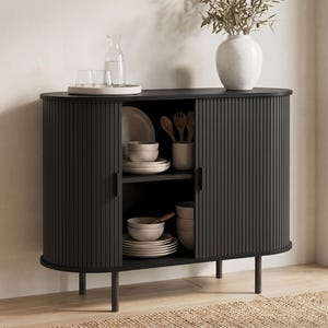 May include: A black cabinet with a ribbed door design, featuring a curved shape and short legs. The cabinet is open, revealing shelves with white dishes, bowls, and wooden utensils. A white vase with greenery and a tray with glasses and a water carafe sit on top.