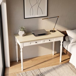 May include: A white wooden desk with a light beige rectangular top and two drawers featuring small dark knobs. The desk has four turned legs and holds a closed gray laptop, a small potted succulent in a geometric gray planter, and a black desk lamp.