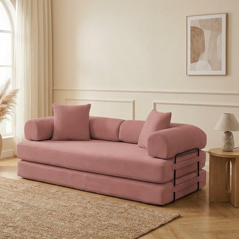 Modern green convertible folding sofa bed with ribbed corduroy fabric in a minimalist living room, featuring modular foam design, removable backrests, matching pillows, beige rug, and soft neutral decor near a large window.