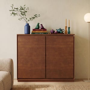 May include: A rectangular wooden cabinet with two doors, topped with a blue vase containing a leafy branch, two bird figurines, three stacked books titled Vanity Fair 100 Years, a purple bowl, a green teapot, and two tall candle holders.