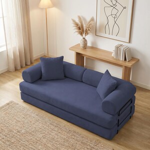 Modern green convertible folding sofa bed with ribbed corduroy fabric in a minimalist living room, featuring modular foam design, removable backrests, matching pillows, beige rug, and soft neutral decor near a large window.