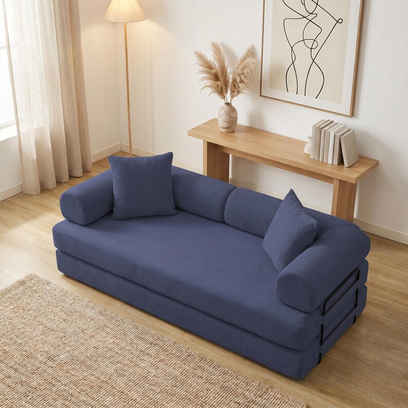 Modern green convertible folding sofa bed with ribbed corduroy fabric in a minimalist living room, featuring modular foam design, removable backrests, matching pillows, beige rug, and soft neutral decor near a large window.
