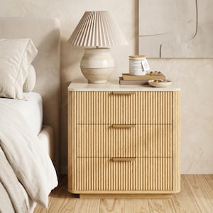May include: A light wood nightstand with three drawers featuring vertical fluted detailing and slim brass-toned handles. The piece has a smooth white top surface and sits on a recessed base, providing a clean look for a bedroom setting.