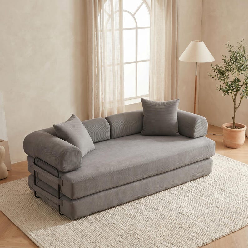 Modern green convertible folding sofa bed with ribbed corduroy fabric in a minimalist living room, featuring modular foam design, removable backrests, matching pillows, beige rug, and soft neutral decor near a large window.
