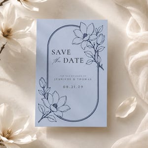May include: A light blue rectangular Save the Date card featuring a dark blue arched frame and line art of magnolia flowers. The text reads: SAVE the DATE, FOR THE WEDDING OF JENNIFER & THOMAS, 08.21.29. The design includes floral accents.