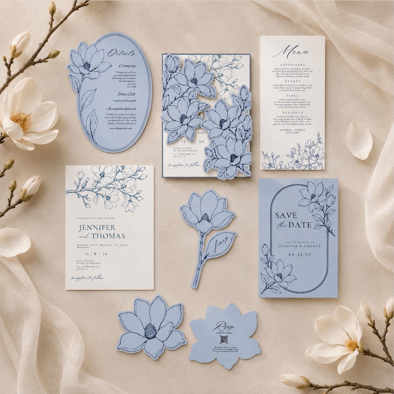 May include: A wedding stationery set featuring light blue and white paper cards with dark blue line art of magnolia flowers. The collection includes a rectangular invitation, a menu, a save the date card, an oval details card, and floral-shaped inserts.