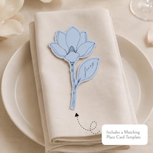 May include: A light blue paper place card shaped like a magnolia flower with a stem and a leaf. The name Jenny is written in dark blue cursive script on the leaf. A small white rectangular box contains the text Includes a Matching Place Card Template.