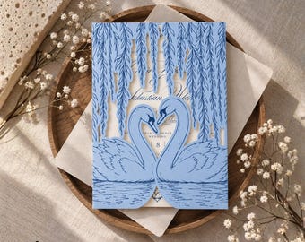 Puede incluir: Una cubierta de invitación de boda azul claro cortada con láser que presenta una ilustración detallada de dos cisnes frente a frente bajo ramas de sauce. El diseño intrincado incluye ondas de agua en la base y texto visible a través de los recortes.