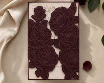 Puede incluir: Una tarjeta de invitación rectangular con una superposición floral de color burdeos oscuro cortada con láser y patrones de rosas intrincados. La tarjeta interior color crema muestra rosas de línea fina y texto. Una rosa burdeos.