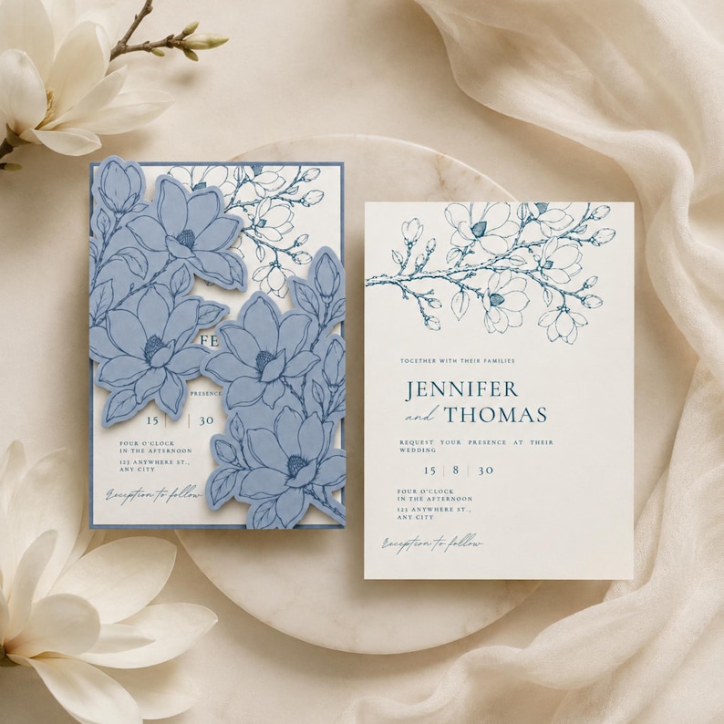May include: A wedding invitation set featuring a white card with blue line-art magnolia illustrations and a matching blue die-cut overlay with layered floral shapes. The text reads Jennifer and Thomas, 15 8 30, four o'clock in the afternoon, 123 anywhere st., any city, and reception to follow.