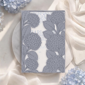 May include: A rectangular invitation card featuring a laser-cut, periwinkle blue paper overlay with intricate hydrangea flower and leaf patterns. The overlay frames a white inner card with visible text including the name Charlotte and the number 30.