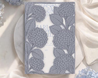 Puede incluir: Una tarjeta de invitación rectangular con una superposición de papel azul bígaro cortada con láser, con intrincados patrones de flores y hojas de hortensia. Enmarca una tarjeta interior blanca con texto visible que incluye el nombre Charlotte y el número 30.