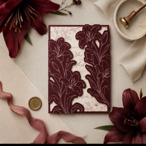 May include: A rectangular wedding invitation card featuring a deep burgundy laser-cut cover with intricate lily flower outlines. The cream-colored inner card displays elegant script text. A gold wax seal and a matching burgundy lily are nearby.