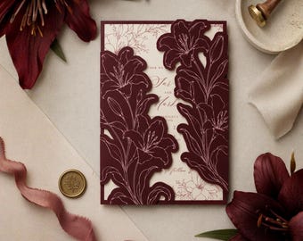 Puede incluir: Una tarjeta de invitación de boda rectangular con una cubierta de color burdeos profundo cortada con láser y contornos de lirios. La tarjeta interior de color crema muestra un texto elegante. Un sello de cera dorado y un lirio burdeos están cerca.
