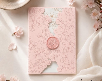 Puede incluir: Una tarjeta de invitación rosa claro con un delicado patrón de líneas de flores de cerezo, asegurada en el centro por un sello de cera circular con un motivo floral. La tarjeta está entreabierta, revelando un panel interior blanco con detalles dorados.