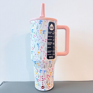 Puede incluir: Un vaso grande y aislado con un estampado floral en rosa, azul y amarillo. Tiene un asa, tapa y pajita rosas. El vaso tiene una capacidad de 1,18 litros y presenta el texto "HydroJug".