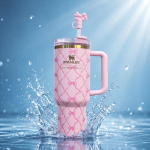 Puede incluir: Un vaso Stanley rosa con asa y pajita, con un dise&ntilde;o de lazo y el texto "LOVESHACKFANCY". El vaso est&aacute; decorado con un patr&oacute;n de lazos rosas y tiene un asa y pajita rosas. El vaso est&aacute; en un chapoteo de agua.