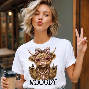May include: White t-shirt featuring a cartoon Highland cow wearing heart-shaped sunglasses, a leopard print bow, and a pearl necklace. The word "MOOODY" is printed below the cow. The person is holding a coffee cup.