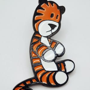 May include: A metal enamel pin shaped like a cartoon tiger sitting down. The tiger features bright orange fur with black stripes, a white face, white chest, and white paws. The design is outlined in black metal, creating a bold, graphic appearance.