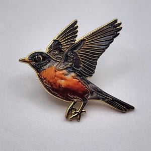 May include: A gold-toned metal enamel pin shaped like a bird in flight. The bird features a dark gray head and wings with detailed feather lines, a bright orange chest, and a small gold beak. The pin is captured in a side profile view.