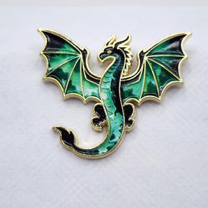 May include: A gold-toned metal enamel pin shaped like a dragon with outstretched wings. The dragon features a gradient of emerald green and dark forest green enamel fill, with a slender body, curved tail, and detailed wing membranes outlined in gold.
