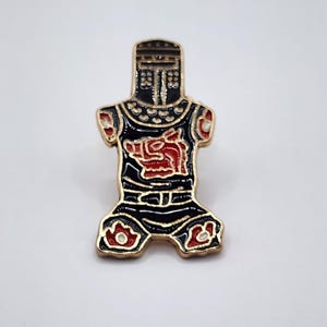 May include: A metal enamel pin shaped like a suit of armor with a helmet, featuring a black base with gold-toned outlines. The chest plate displays a red boar emblem, and the lower section includes red and white decorative accents on the armor.