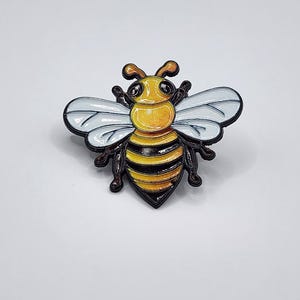 May include: A bee-shaped enamel pin featuring a yellow and black striped abdomen, a yellow head with black eyes, and two translucent white wings with black outlines. The insect design has a glossy finish and is rendered in a cartoon style.