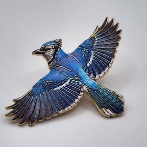 May include: A blue jay bird pin featuring vibrant blue, white, and black enamel detailing with gold-toned metal edges. The bird is depicted in a flying position with wings spread wide, showcasing intricate feather textures and a pointed beak.