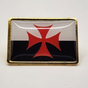 May include: A rectangular metal pin with a gold-toned border featuring a glossy finish. The design displays a horizontal bicolor flag with a white upper half and a black lower half, centered with a bold red cross pattée symbol.