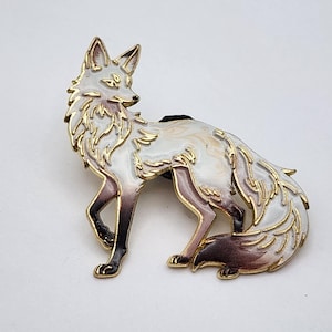 May include: A metal enamel pin shaped like a fox with white fur, gold-toned outlines, and dark brown shading on the legs and tail. The fox is depicted in a standing position with its head turned to the side, featuring intricate detailing.