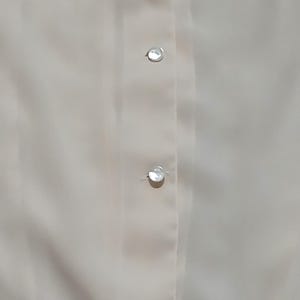 Könnte beinhalten: Nahaufnahme einer cremefarbenen Bluse mit drei durchsichtigen, irisierenden Knöpfen auf der Vorderseite. Die Knöpfe sind rund und scheinen aus einem reflektierenden Material zu bestehen. Der Stoff hat eine glatte Textur.
