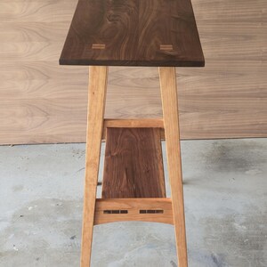 Puede incluir: Una mesa auxiliar de madera hecha a mano con una tapa rectangular marr&oacute;n oscuro y patas de color marr&oacute;n claro. La mesa presenta un estante inferior y ensamblajes decorativos. La mesa mide aproximadamente 76 cm de altura.
