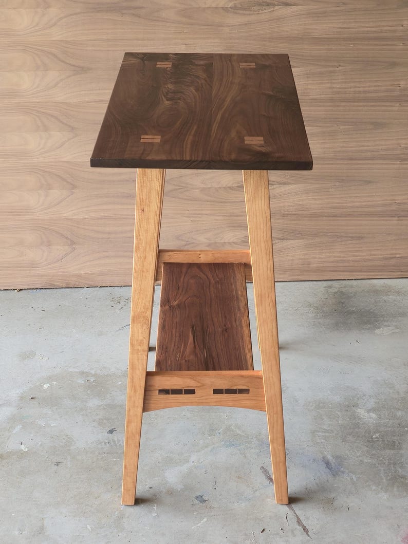 Puede incluir: Una mesa auxiliar de madera hecha a mano con una tapa rectangular marr&oacute;n oscuro y patas de color marr&oacute;n claro. La mesa presenta un estante inferior y ensamblajes decorativos. La mesa mide aproximadamente 76 cm de altura.