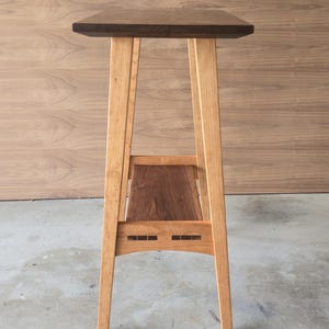 Puede incluir: Un soporte de madera alto para plantas con una parte superior rectangular de color marr&oacute;n oscuro y un estante inferior. Las patas son de madera de color claro y el estante es de madera m&aacute;s oscura. El soporte mide aproximadamente 76 cm de alto.