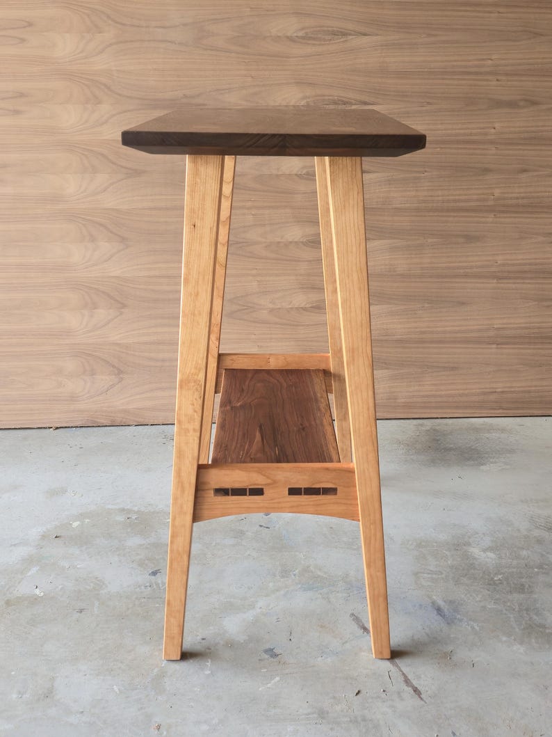 Puede incluir: Un soporte de madera alto para plantas con una parte superior rectangular de color marr&oacute;n oscuro y un estante inferior. Las patas son de madera de color claro y el estante es de madera m&aacute;s oscura. El soporte mide aproximadamente 76 cm de alto.
